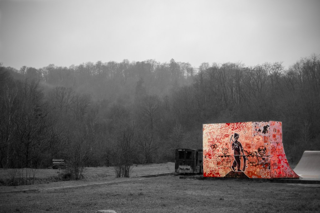 A dark and grayish landscape with forest in the background. In the foreground in bright, vivid colors is a wall which is part of a skatepark.