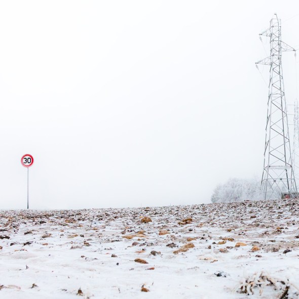 A lonely speed sign stands in an empty field. On the right side there are electricity pylons. The field is covered with thin layer of snow. The background is empty and quite foggy. The ambiance is frosty and full of desolation.