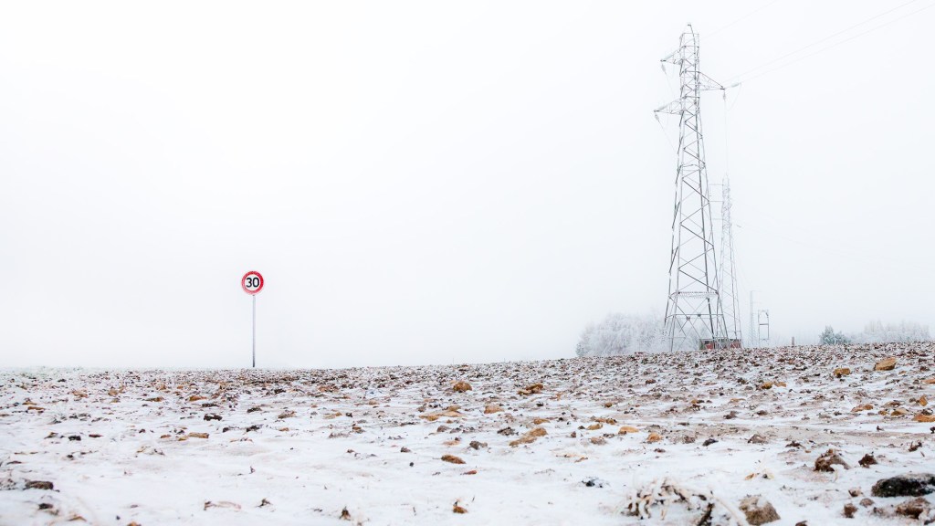 A lonely speed sign stands in an empty field. On the right side there are electricity pylons. The field is covered with thin layer of snow. The background is empty and quite foggy. The ambiance is frosty and full of desolation.